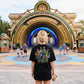 May include: Black t-shirt with a colorful graphic design on the back, featuring various characters. The shirt is worn by a person standing in front of an archway with the text "YOUR DREAMS ARE BORN WHERE THE IMPOSSIBLE LIVES."